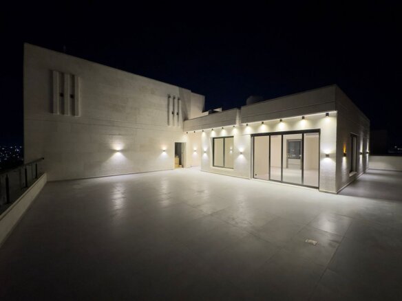 Nighttime rooftop terrace of a modern building with warm wall lights and large sliding glass doors, beige stone walls. An open doorway and city lights in the distance.