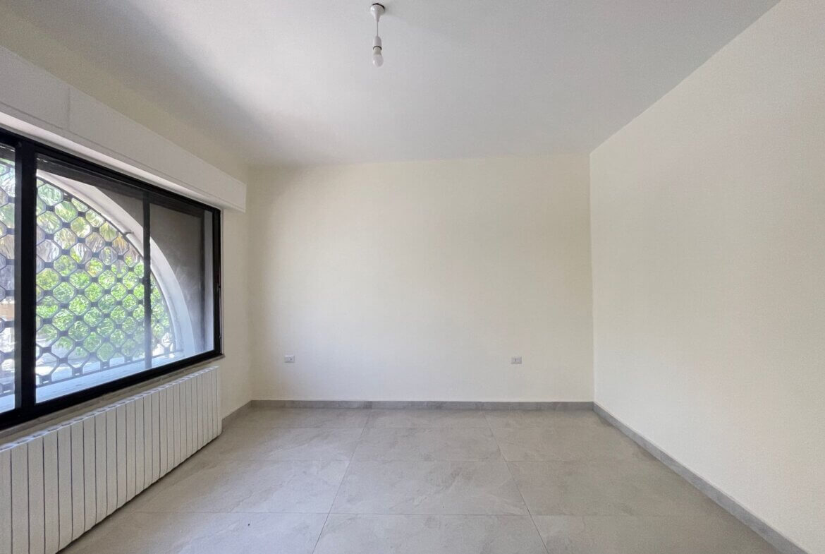 7101002c-51c6-4f76-898d-df2df0380197 Empty, unfurnished room with a large left-side window featuring a metal hexagonal grille, white walls, and light gray tiled floor; a radiator runs along the window wall and a single bare light bulb hangs from the ceiling.
