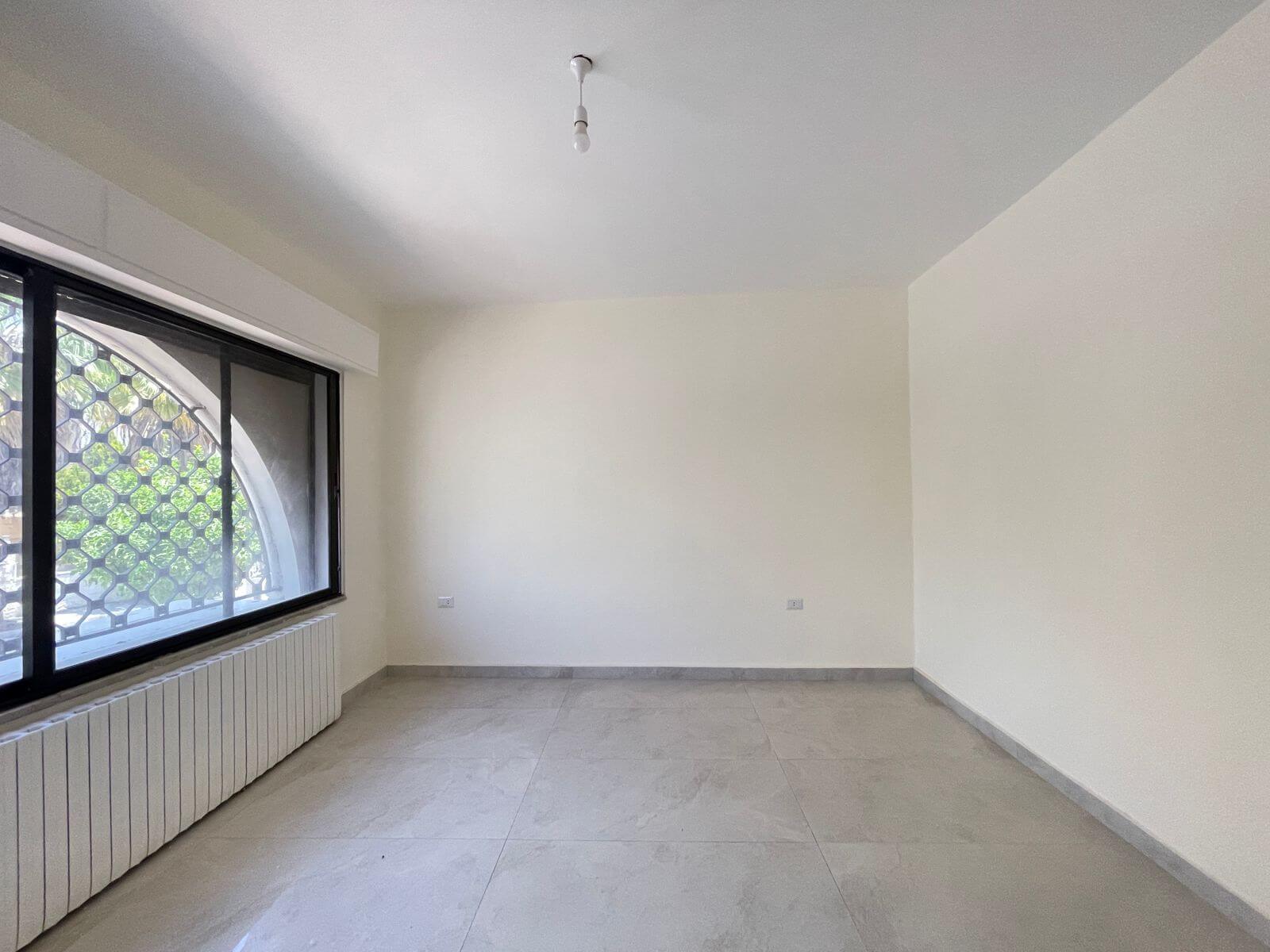 7101002c-51c6-4f76-898d-df2df0380197 Empty, unfurnished room with a large left-side window featuring a metal hexagonal grille, white walls, and light gray tiled floor; a radiator runs along the window wall and a single bare light bulb hangs from the ceiling.