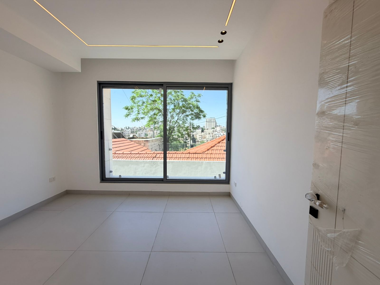 714c4166-a250-4535-b363-3bbeef4440fd Empty white room with a large sliding window overlooking red-tiled rooftops and a cityscape outside