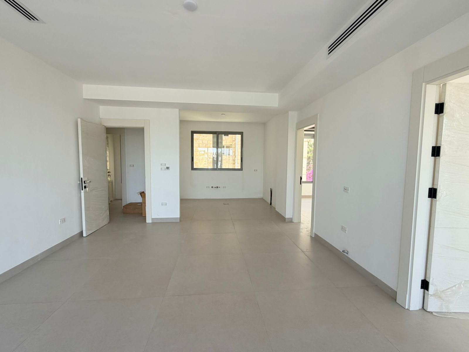 72599319-0d64-4a55-bd38-933e13f28cee Bright empty living area with white walls, large central window, and light gray tiled floor; multiple doorways lead to other rooms in a modern apartment.