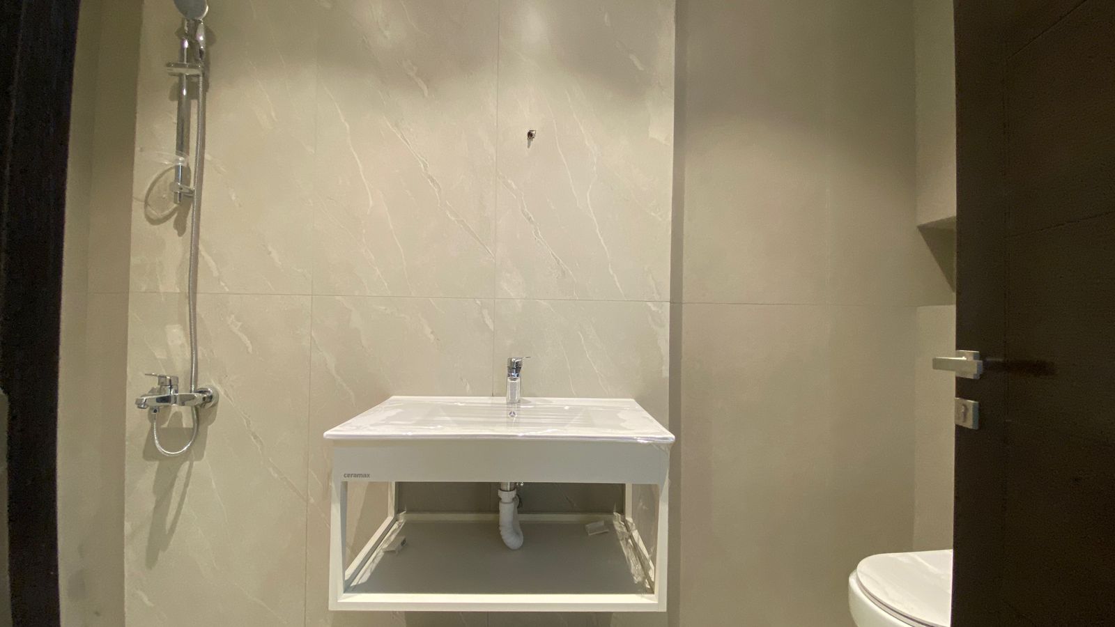 732ec3df-28a6-48a5-84cd-1cd4ad45f0aa Small modern bathroom with a white wall-mounted sink, exposed pipes underneath, and a handheld shower to the left; beige marble-tiled walls and a dark door on the right.
