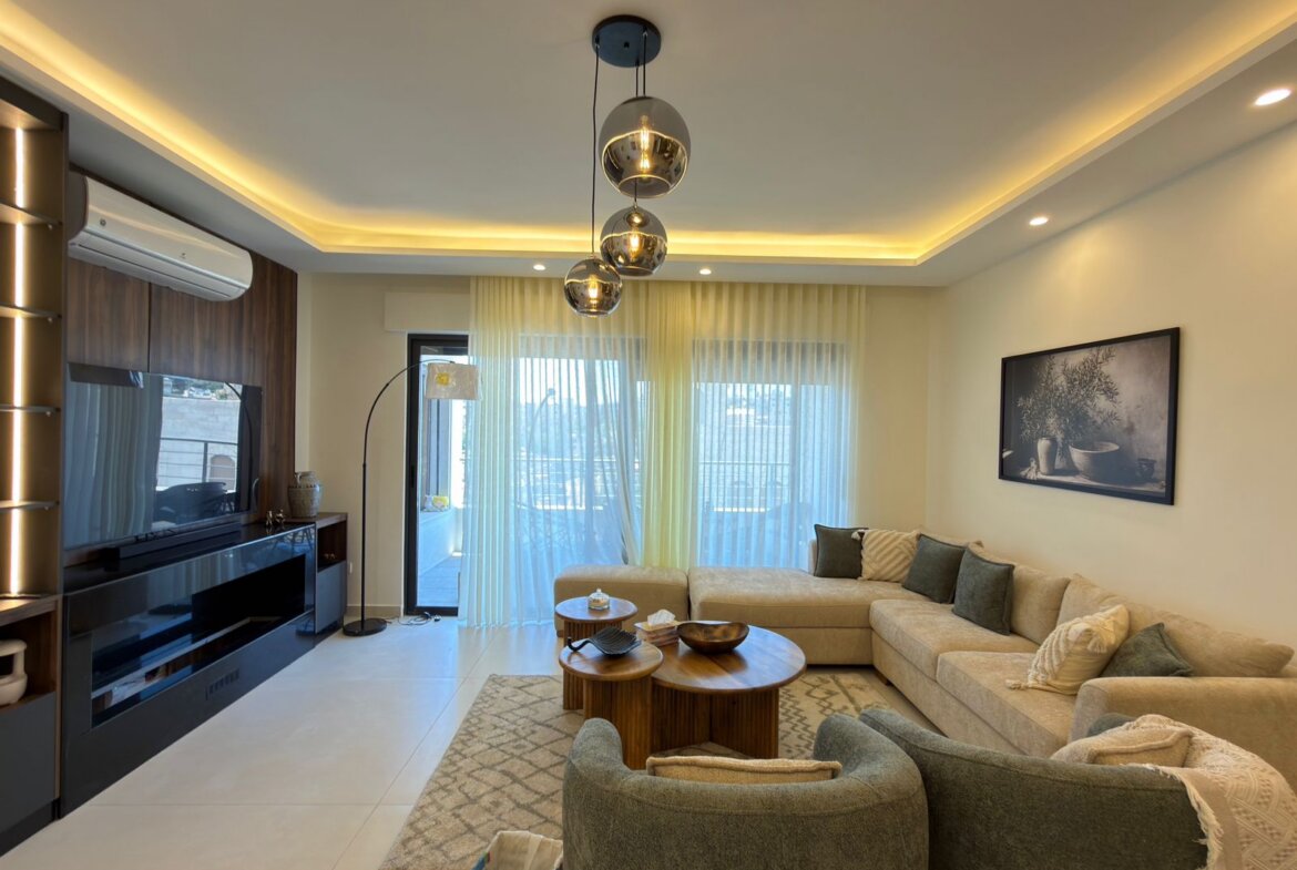 Modern living room with a beige sectional, round wooden coffee tables, and a textured rug under soft lighting from ceiling pendants and LED strips.