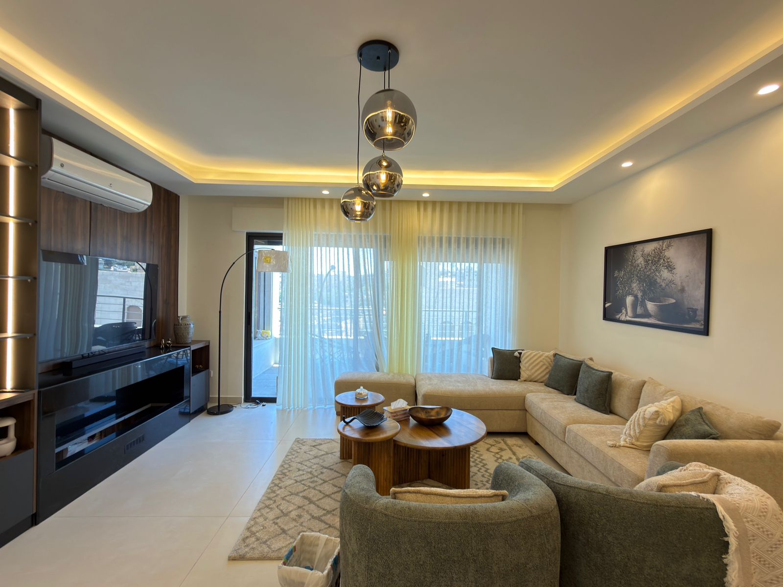 73b9ca44-079c-49f2-b003-c3ef034649ce Modern living room with a beige sectional, round wooden coffee tables, and a textured rug under soft lighting from ceiling pendants and LED strips.