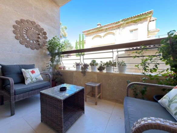 Cozy balcony seating area with wicker furniture, cushions, and potted plants along the railing under a bright blue sky.