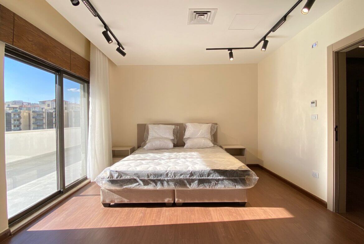 Bright modern bedroom with a double bed in the center, plastic-covered mattress, beige walls, and a large glass door opening to a balcony on the left letting in sunlight.
