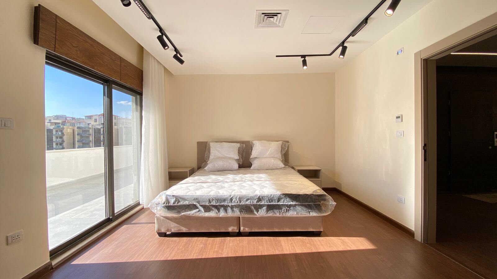 Bright modern bedroom with a double bed in the center, plastic-covered mattress, beige walls, and a large glass door opening to a balcony on the left letting in sunlight.