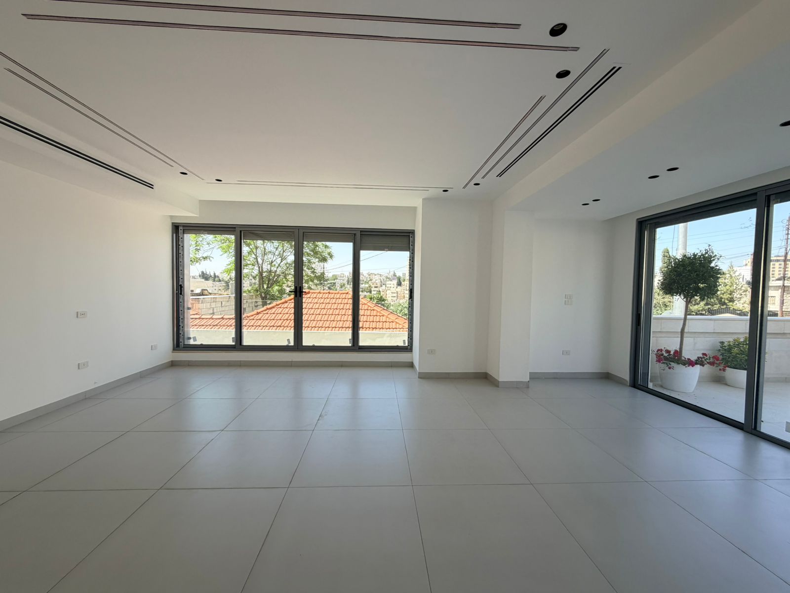 7404cd03-dbee-4cef-adbf-aade802bf237 Bright empty living room with large glass doors to a balcony, white walls, gray tiled floor, and decorative ceiling lines.