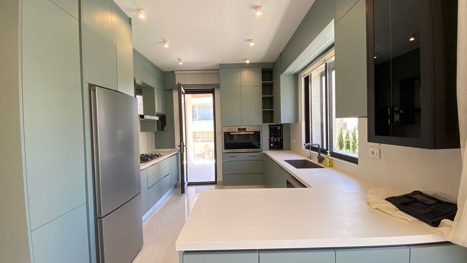 753aeb08-ca93-401a-b481-963efa9df872 Modern kitchen with pale green cabinets, white countertops, and stainless steel appliances bathed in natural light from a large window/door.