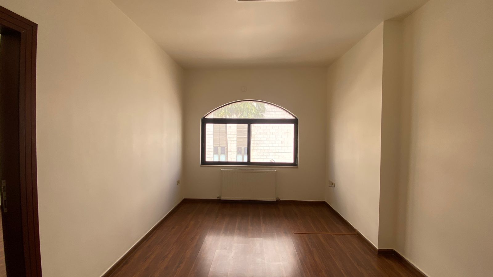 759c08eb-42e9-4814-8b8e-9c48a5d66966 Empty rectangular room with a curved-top window, radiator under the window, and brown wood floor.
