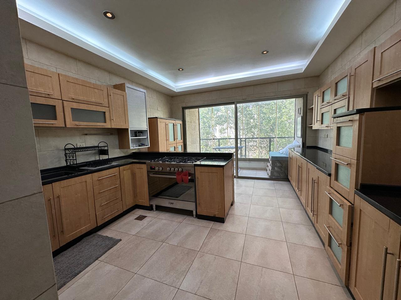 76673674-5564-4e3d-abaa-2d294de133b5 Modern kitchen with light wood cabinets, black countertops, and a central island leading to a balcony door.