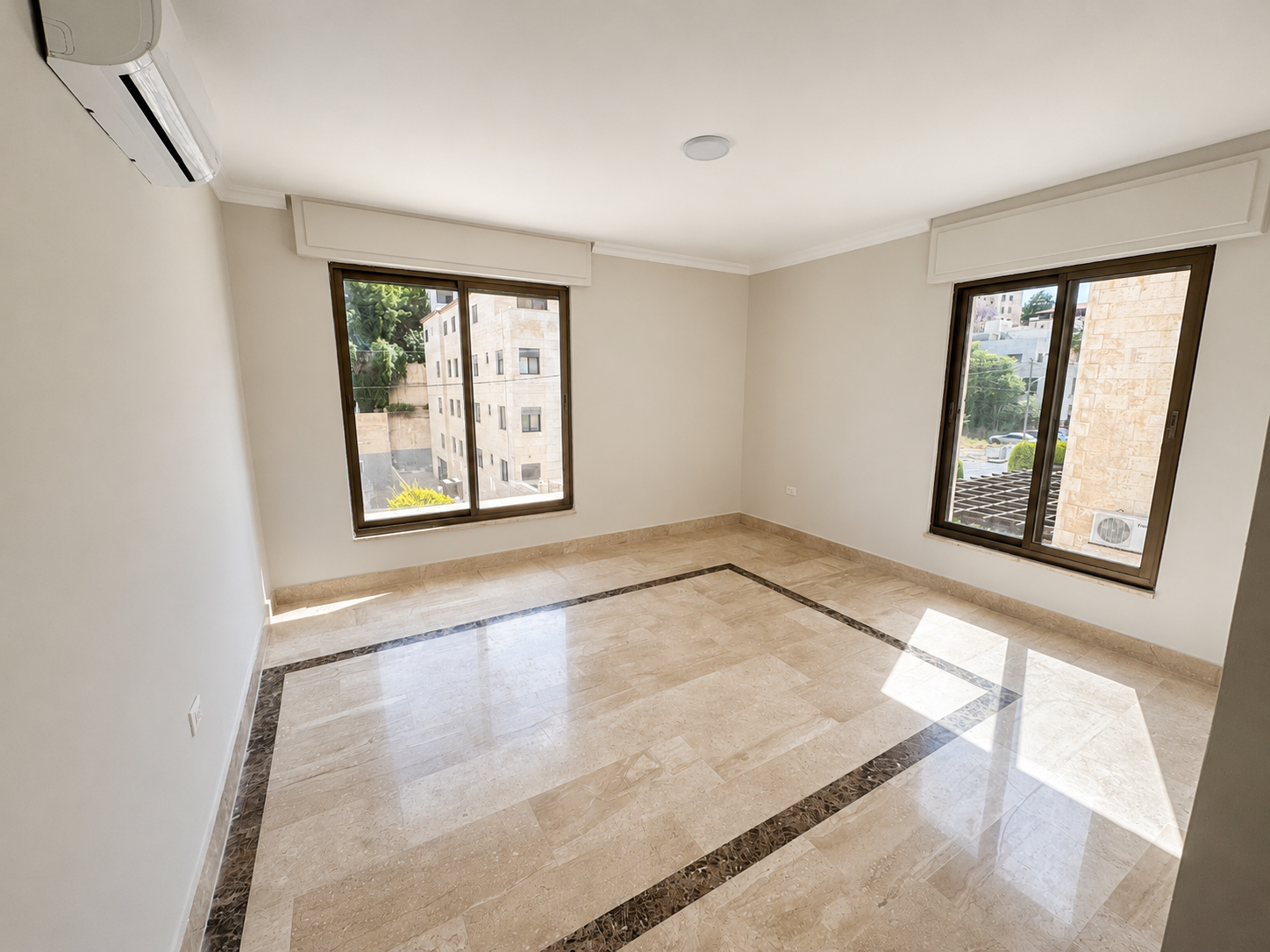 7677ca34-c136-4f93-8c6e-39d9ca26e341 Sunlit empty living room with beige walls, marble floor with dark border, and two large windows showing buildings outside; wall AC unit mounted high left.