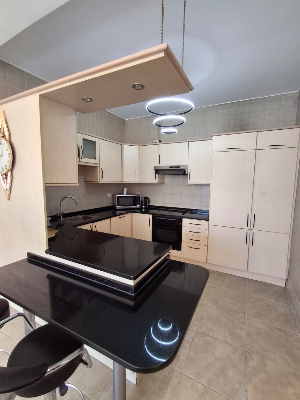 79078298-7f26-4cb7-9068-26e541b3a9f4 Modern L-shaped kitchen with beige cabinets and a glossy black island under circular pendant lights above.