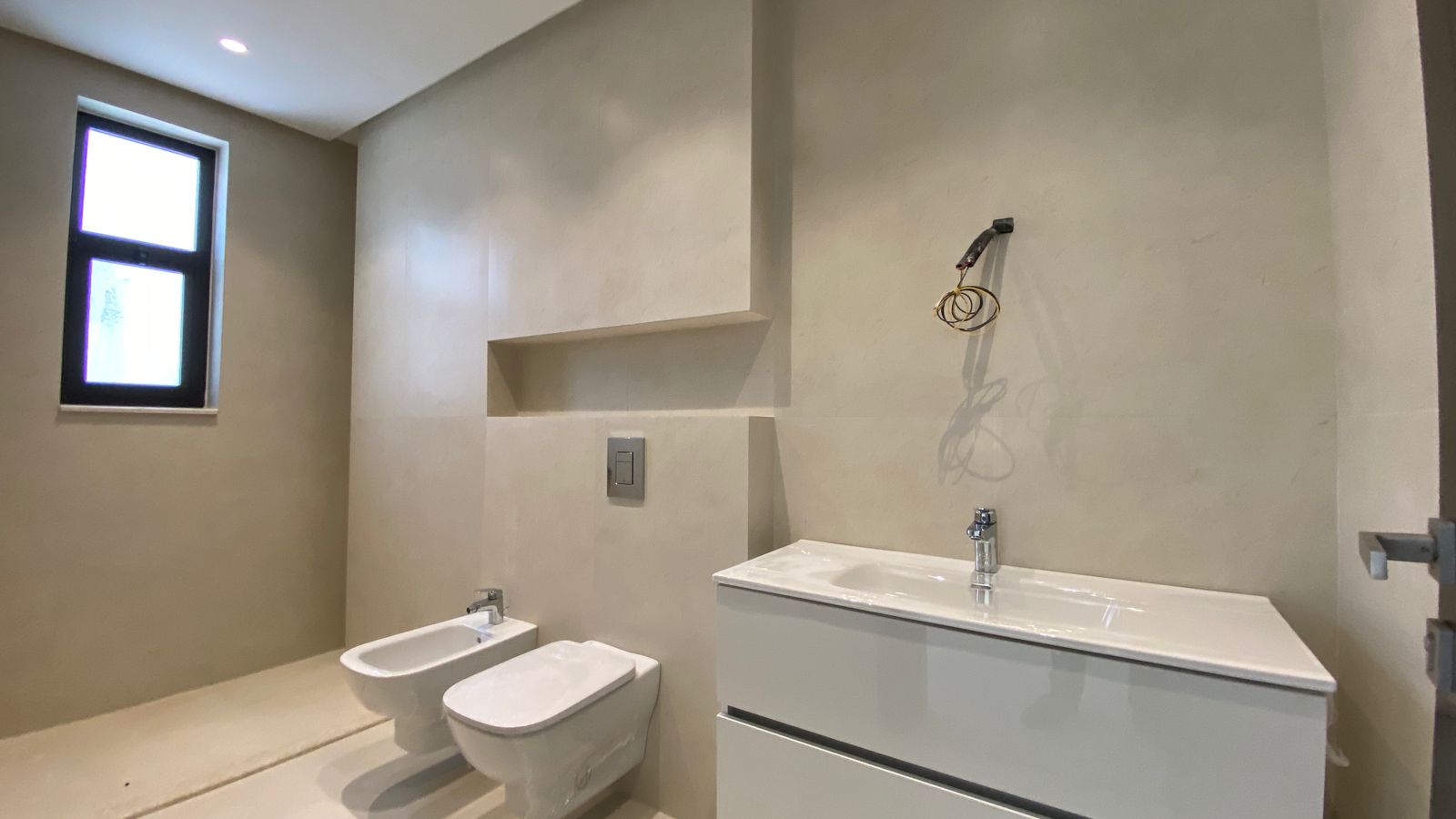 79317639-37ee-4879-9ac8-8380195448eb Unfinished modern bathroom with wall-mounted toilet and bidet, white vanity, and exposed wiring near the sink area.