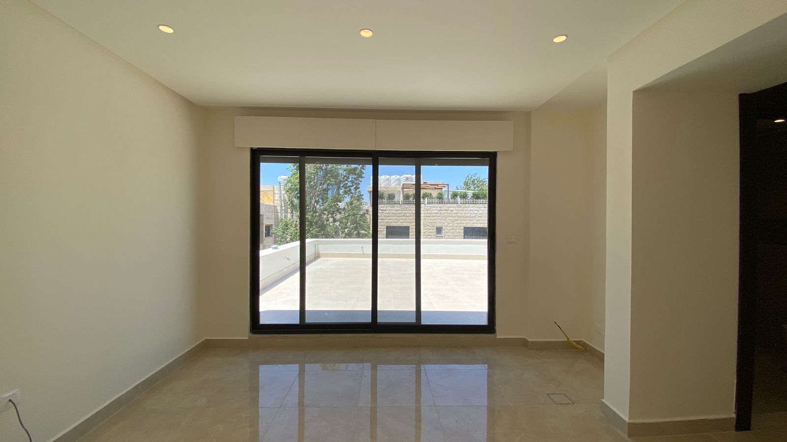 7982c041-c9f1-4c38-a113-56381d53c35b Bright empty living room with black-framed sliding glass doors opening to a sunlit patio outside a brick wall.