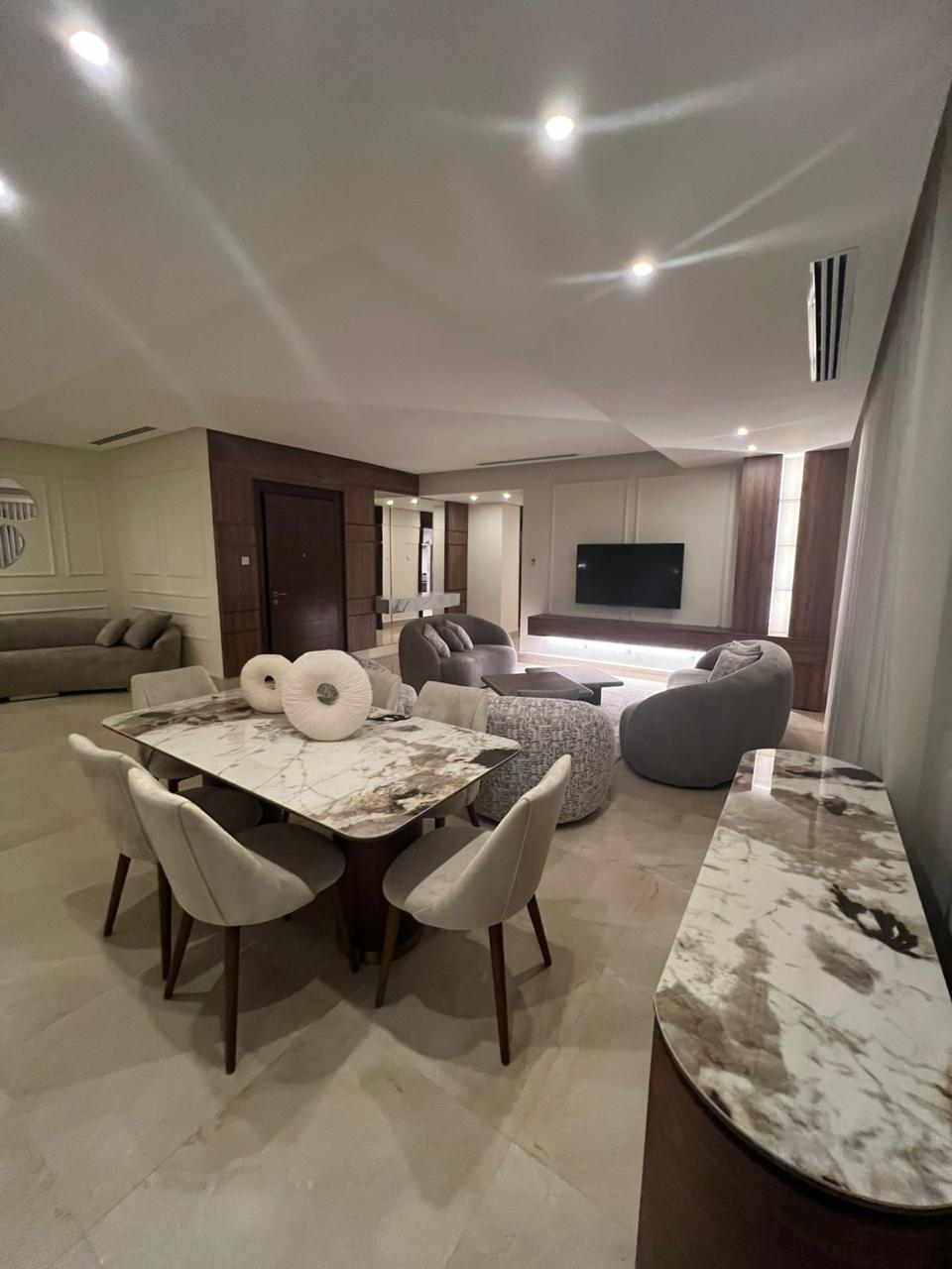 7a7895a2-9fdf-46e4-b46e-06c4b5813e9d Open-plan living and dining area with a marble dining table surrounded by beige upholstered chairs, a gray lounge area, and a wall-mounted TV.