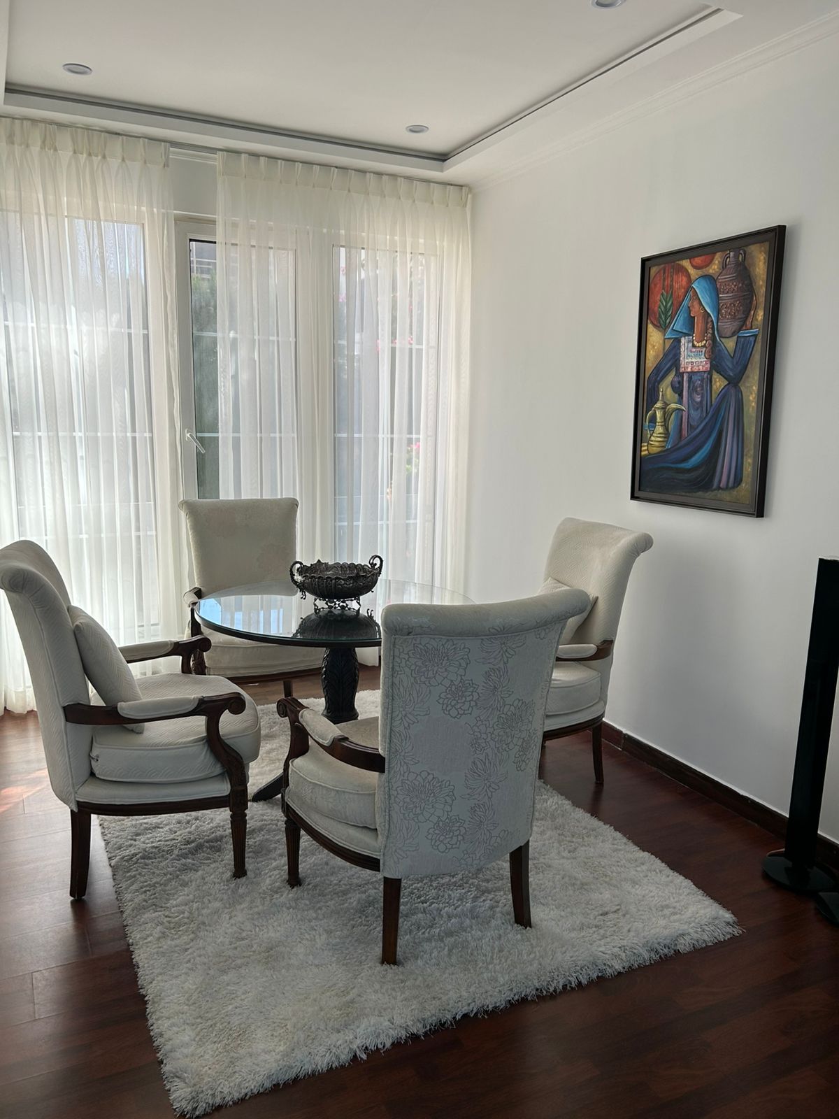 7afc437e-b656-45ef-b9c6-508cba9ed56a Bright living/dining area with a round glass table and four upholstered cream chairs on a white shag rug, sheer curtains, and a colorful wall painting.