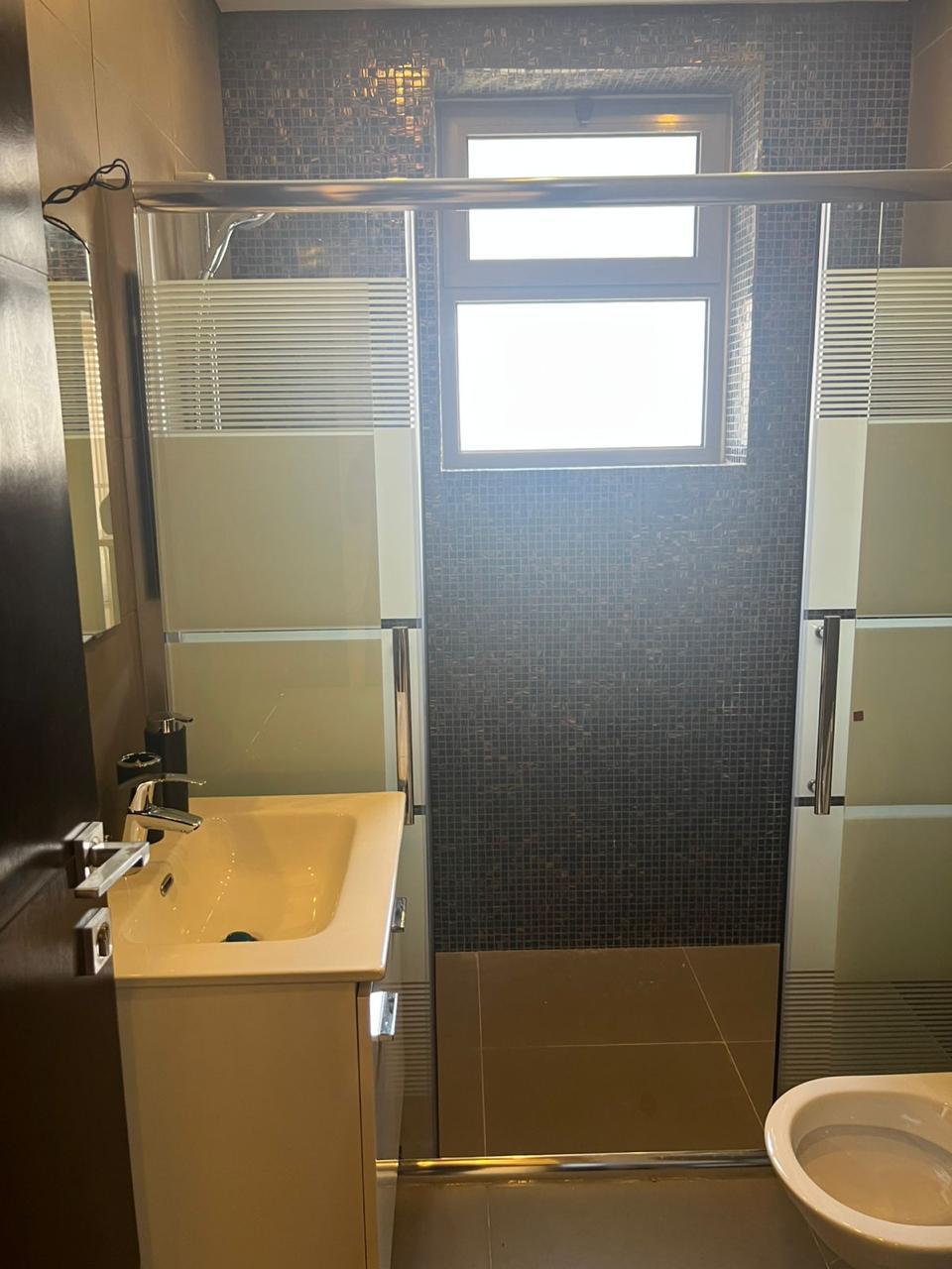 7cd9d4c4-6c0b-4ba9-afd1-09a959d8dee4 Modern bathroom with glass shower enclosure featuring frosted panels, dark mosaic tiled wall, and a rectangular window above the shower.