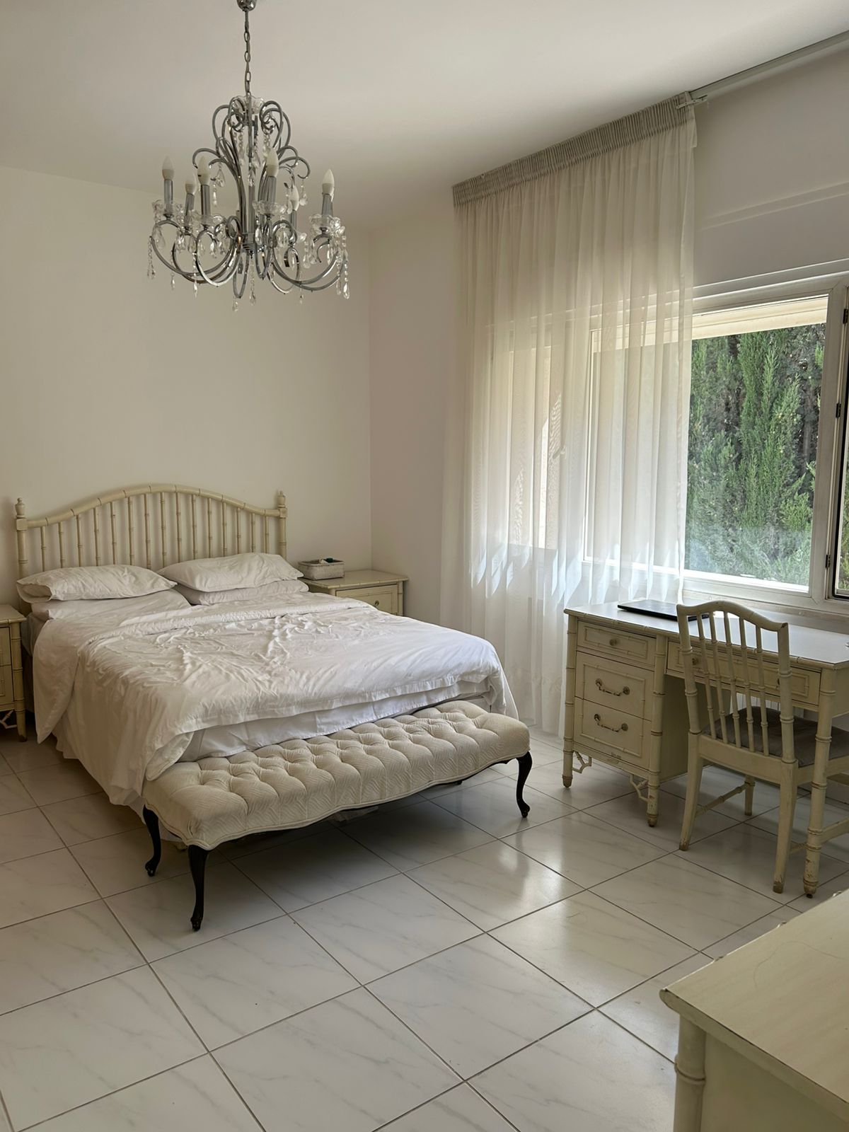 7ce8f9f5-464c-46be-9ea6-f09e74a6a23d Bright white bedroom featuring a metal bed frame, white bedding, a tufted ottoman at the foot, and a chandelier hanging from the ceiling.