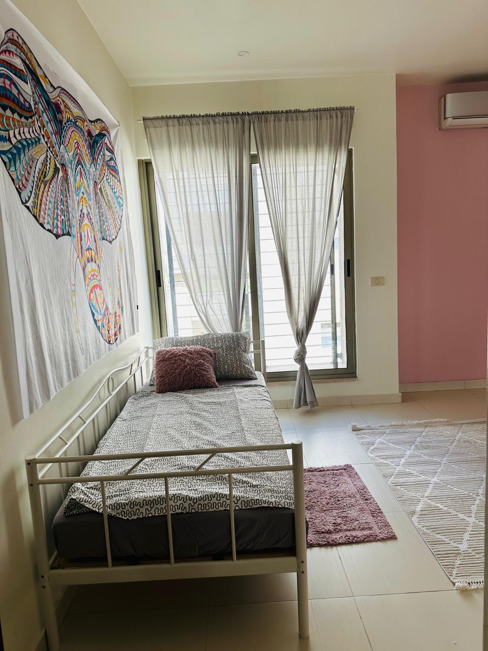 7d2ed4d9-bcc8-449b-a6fd-eb7a8ab302c7 Single white metal bed with gray geometric bedding, two pillows, and a pink fuzzy cushion under a large window with sheer gray curtains.