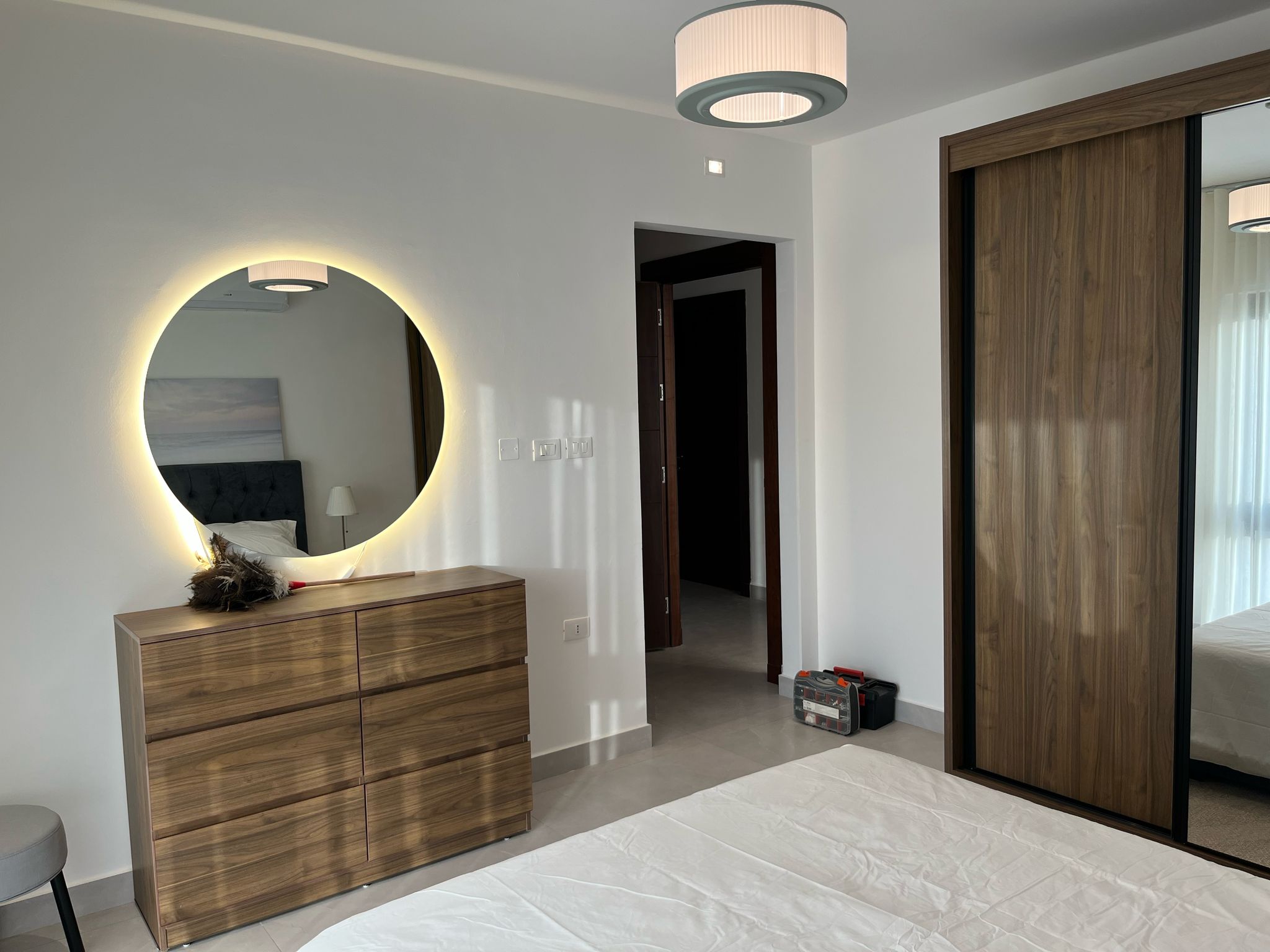 7dd5ca63-709f-4e55-b71f-c23e53ee69ec Bedroom with a round backlit mirror above a wooden dresser, reflecting part of the bed and lamp.