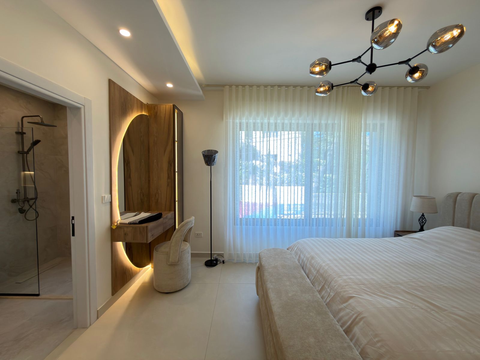 7dd79a5e-b6fd-41c0-9a2e-22a6620f9441 Bright modern hotel bedroom with a large bed, sheer curtains, and a circular-backlit vanity mirror by a wooden desk left of center.