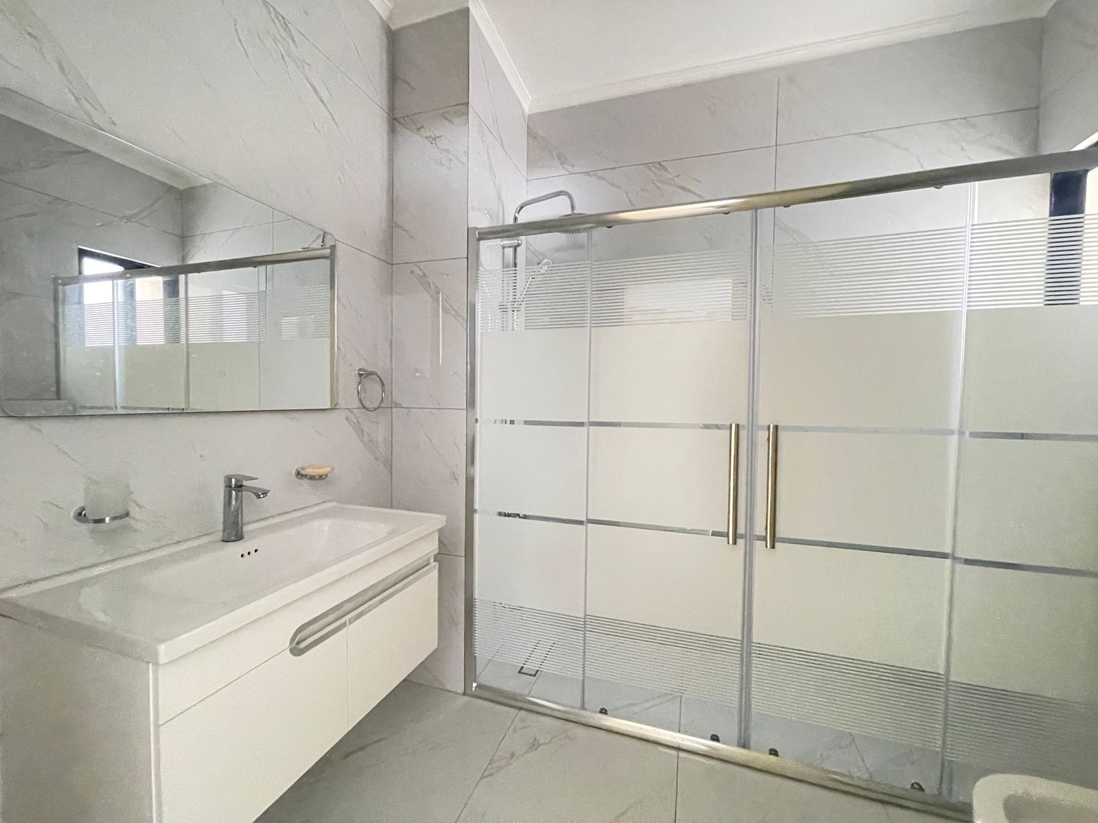7e298767-9a9a-4ab1-b784-3f455d8897dd Modern bathroom with a glass shower enclosure, frosted horizontal bands, and brass handles; white vanity with sink and large mirror above it.