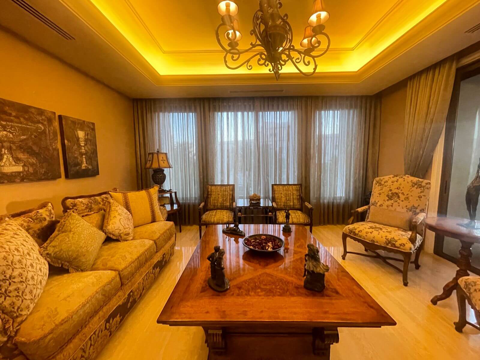 7e61f1d1-bc28-456b-bf8e-4882c266c522 Luxurious living room with an ornate chandelier, beige floral sofa, and patterned armchairs facing a wooden coffee table.