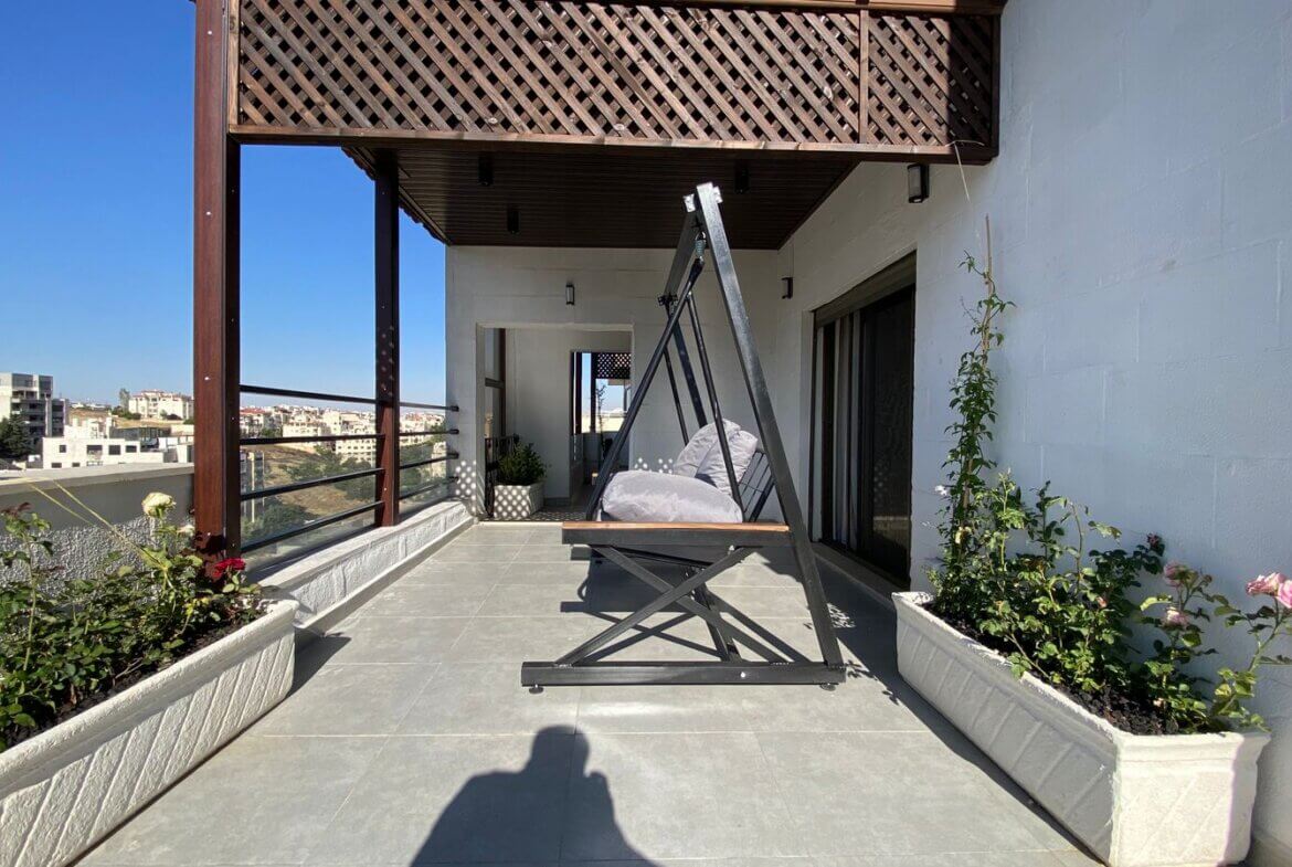 Rooftop patio with a black metal A-frame swing and gray cushions, flanked by white planter boxes and a blue-sky city view.