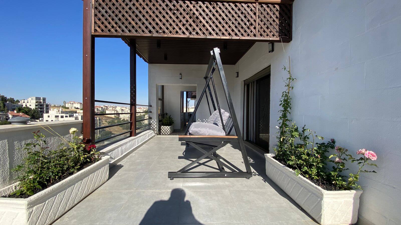 Rooftop patio with a black metal A-frame swing and gray cushions, flanked by white planter boxes and a blue-sky city view.
