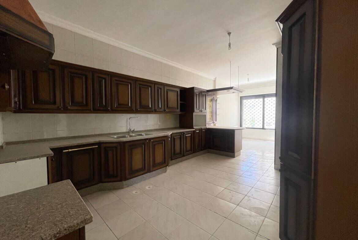 7ff03b24-3edd-4543-8e01-b729e269c3e2 Empty kitchen with dark wood cabinets, white tile backsplash, and a central island near a large window with a grille.