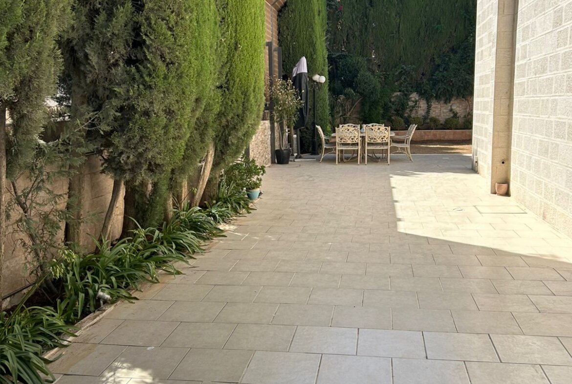 Stone-paved courtyard flanked by tall evergreen hedges, with a distant patio dining table and chairs under a dark umbrella.