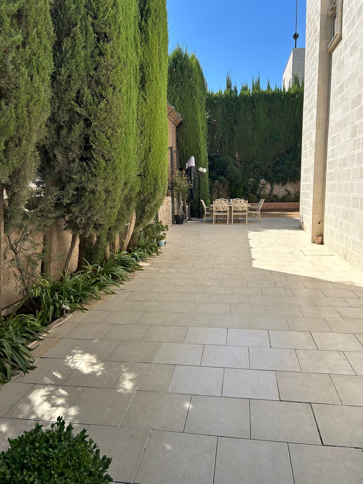 80212b19-1360-4a7d-92d4-883e66a33870 Stone-paved courtyard flanked by tall evergreen hedges, with a distant patio dining table and chairs under a dark umbrella.