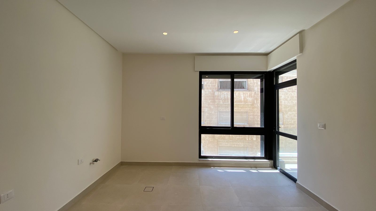 803dcddb-d35f-4967-8a04-3603009cc1bf Empty modern living room with a large black-framed window and glass door to a balcony, beige tile floor, and white walls.