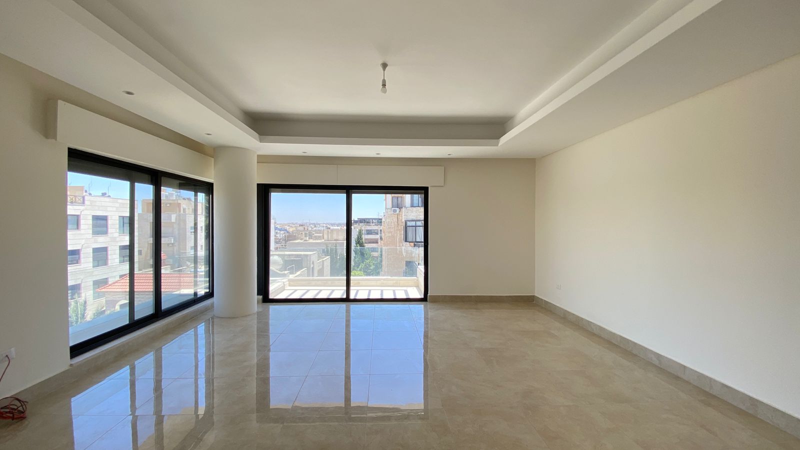 80a6a6eb-ec75-4458-bf14-f979f076f7e1 Bright empty living room with large glass doors to a balcony and city views; beige tile floor and white walls.