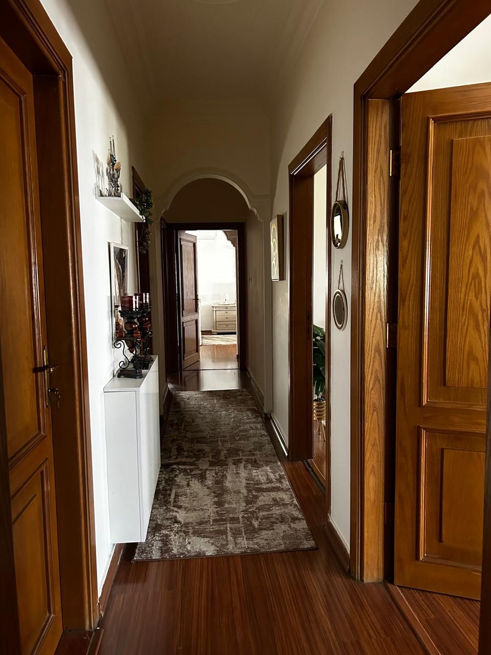 80de9171-482c-42ec-bf34-2f2c50216817 Narrow home hallway with wooden doors on both sides, a patterned rug, and an arched doorway at the far end.