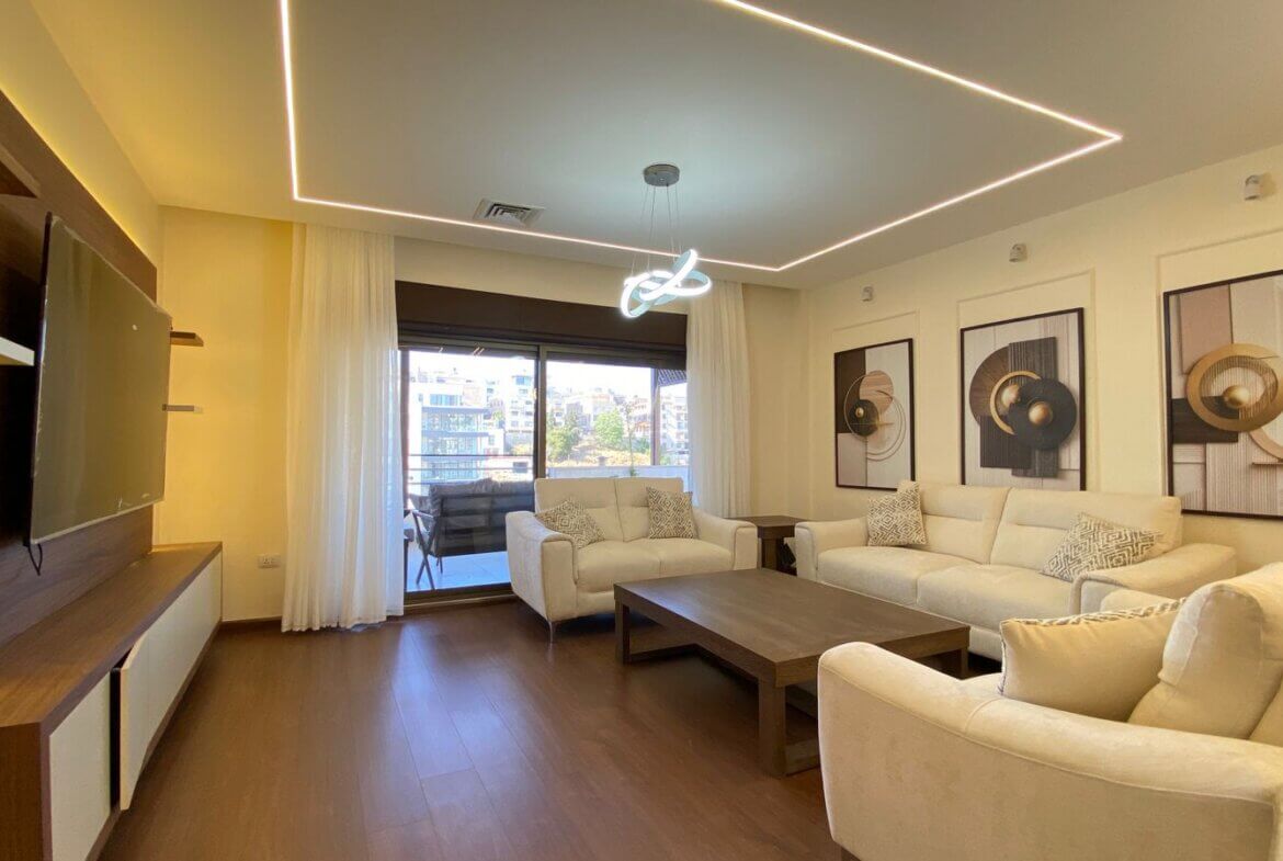 Bright modern living room with white sofas, abstract framed art, and a geometric ceiling light; balcony door with sheer curtains.