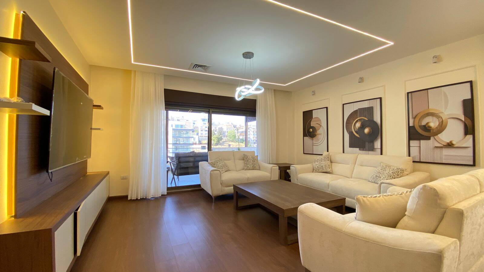 Bright modern living room with white sofas, abstract framed art, and a geometric ceiling light; balcony door with sheer curtains.