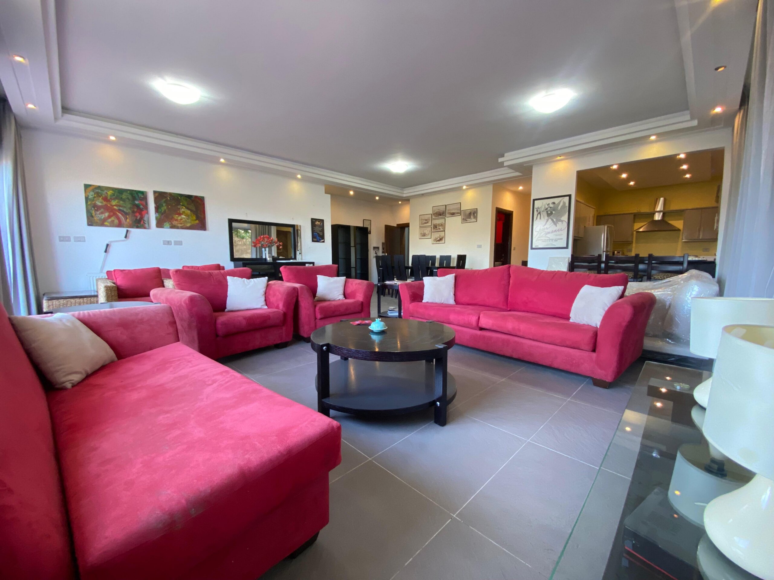 816ecca5-0e29-4b9e-98f1-69fd0de01ab5 Open-plan living room with bright pink sectional sofas, white walls, and a dining area in the background.