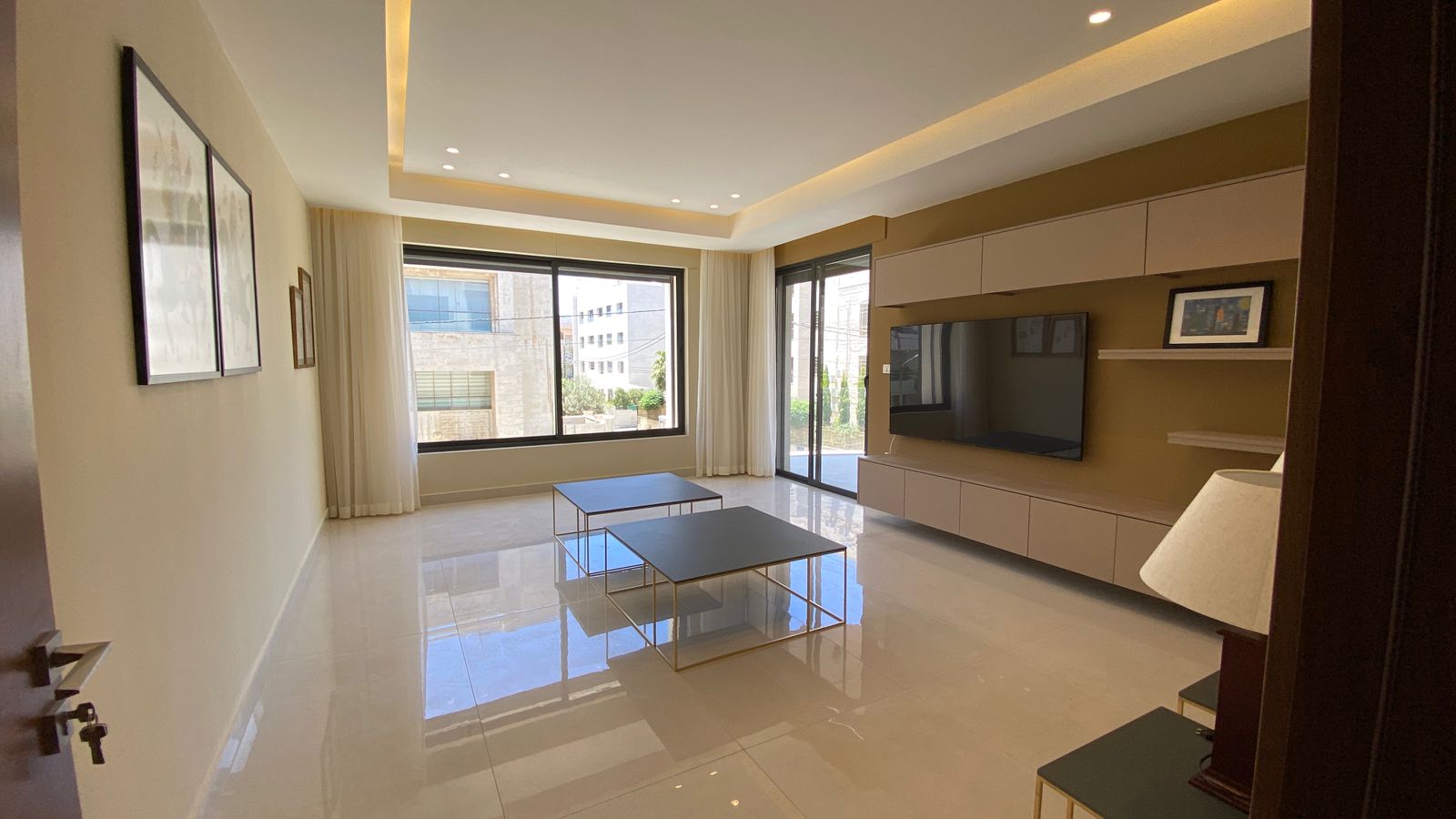 81701bd9-a93b-4234-a5a5-99e17fed58d9 Bright, modern living room with large windows, cream curtains, and two square coffee tables on a glossy tile floor when facing a wall-mounted TV unit.