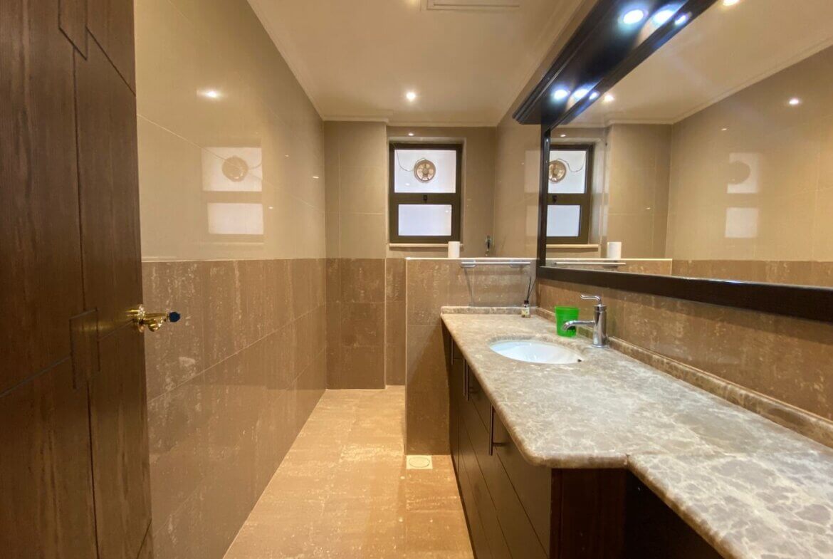 832caf19-4e0e-4834-8815-a6898d654a6c Modern bathroom with marble countertop and sink on the right, dark wood vanity, large mirror, beige tile walls, and a window at the far end.