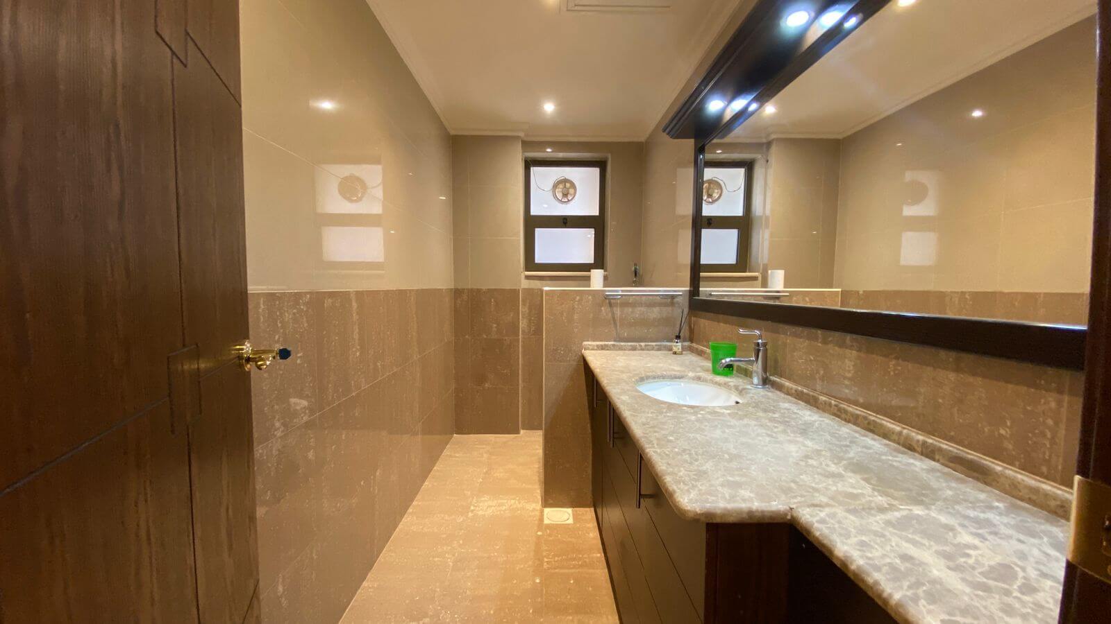 832caf19-4e0e-4834-8815-a6898d654a6c Modern bathroom with marble countertop and sink on the right, dark wood vanity, large mirror, beige tile walls, and a window at the far end.