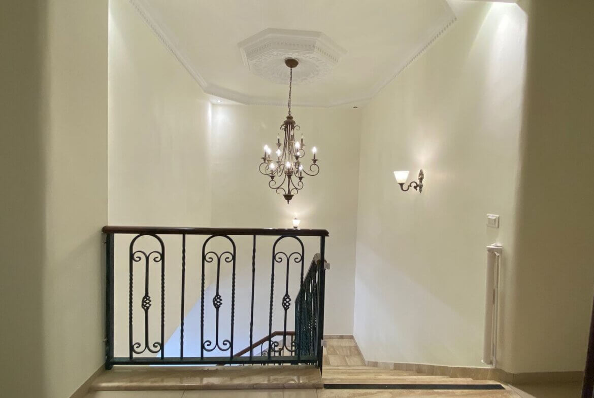 836961f0-a404-4c7b-9cfb-429eb53c14a6 Elegant staircase landing with a decorative chandelier hanging from a coffered ceiling and a wrought-iron railing, in a cream-colored hallway.