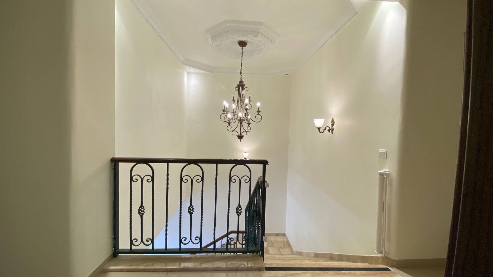 836961f0-a404-4c7b-9cfb-429eb53c14a6 Elegant staircase landing with a decorative chandelier hanging from a coffered ceiling and a wrought-iron railing, in a cream-colored hallway.",