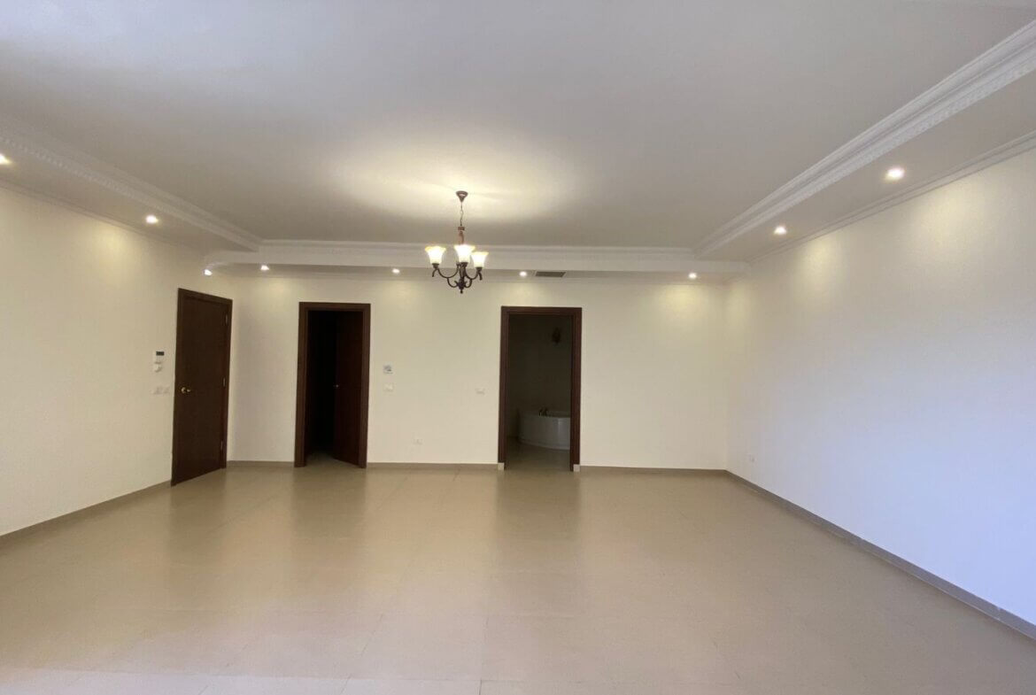 840b3dfa-0f98-4036-8cb8-a30d4cb68559 Empty living room with white walls, beige tile floor, a central chandelier, and three doorways along the back wall, one leading to a bathroom.