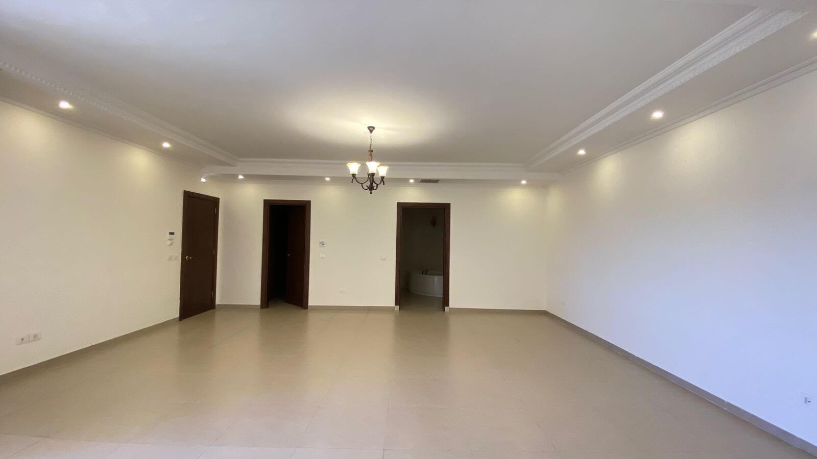 840b3dfa-0f98-4036-8cb8-a30d4cb68559 Empty living room with white walls, beige tile floor, a central chandelier, and three doorways along the back wall, one leading to a bathroom.