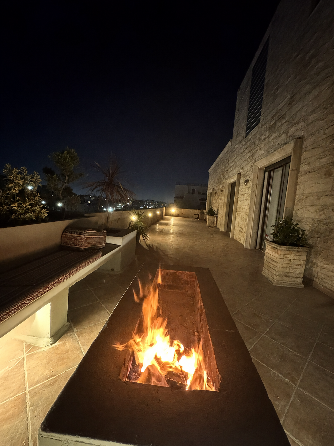 850a0ad0-6876-4075-b3f0-0499d6f4d253 Night-time terrace with a rectangular fire pit in the foreground, beige stone walls, cushioned seating along a low wall, and city lights in the distance.