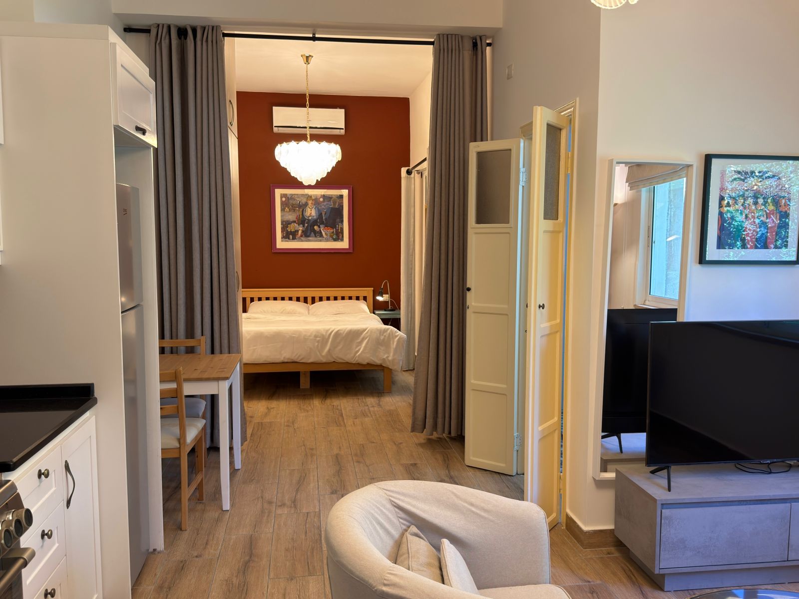 858e38aa-1f1a-4880-9f30-02d4d49bbd34 Bedroom area seen through gray curtains: double bed against a red accent wall with a chandelier above and a framed painting; side table lamp nearby.