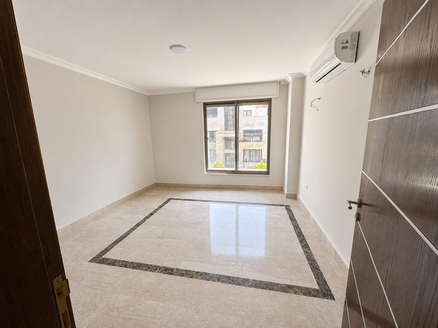 86393a4d-a740-4a35-9065-80e5f03258c8 Empty, sunlit room with beige walls, marble floor, and a large window looking onto a building exterior; wall-mounted air conditioner above the window.