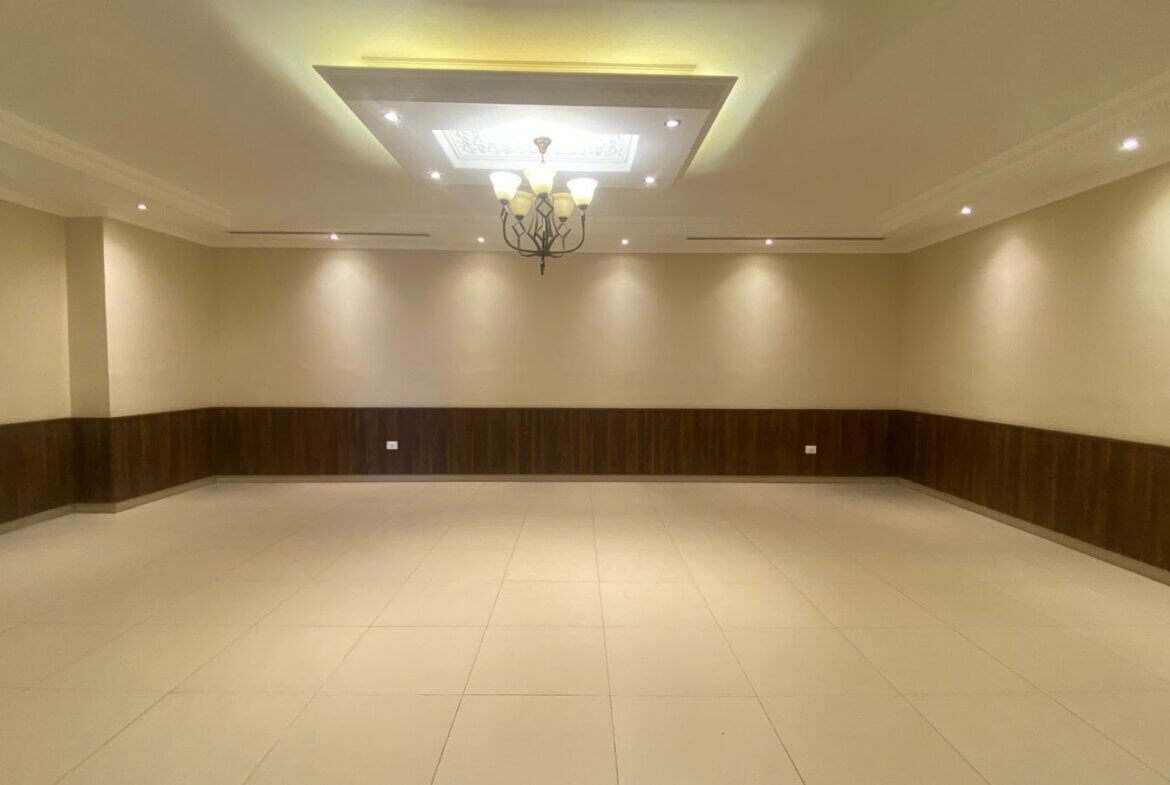 869c197f-7a76-4a8c-b4a3-51721d9e2a91 Empty banquet hall with cream walls, dark wood wainscoting, and a tiled floor under a chandelier.